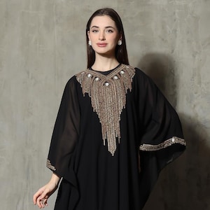 Puede incluir: Un vestido negro estilo caftán con adornos dorados y perlados en el cuello y las mangas. El vestido tiene una silueta fluida y está hecho de una tela transparente. El texto "CAITAN YOSIKA SIKA" es visible en la esquina inferior izquierda.