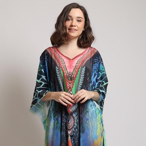 Puede incluir: Un colorido vestido caftán con escote en V y mangas fluidas. El vestido presenta una vibrante mezcla de tonos azules, rojos, naranjas y verdes con un diseño estampado. El texto "CAFTAN YOSIKA" es visible en el lado izquierdo de la imagen.