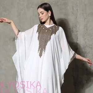 Pode incluir: Um caftan branco esvoaçante com detalhes de contas douradas ao redor do decote e das mangas. O caftan tem mangas largas e uma bainha até o chão. A modelo usa sapatilhas pretas. O texto "CAFTAN YOSIKA" é visível.