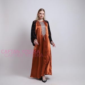 May include: An orange satin caftan with black lace sleeves. The caftan has a decorative silver and gold design down the front. The text "CAFTAN YOSIKA" is visible in the lower left corner.