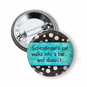 May include: A round button pin with a metal back and pin. The front has a black background with white polka dots and a teal blue stripe with the text "Schrödinger's cat walks into a bar... and doesn't."