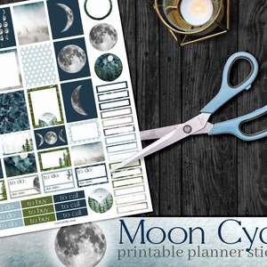 Op de afbeelding: Vel printable planner stickers met een maancyclus thema. De stickers tonen maanfasen, bosscènes en geometrische patronen in blauw, groen en wit. Een schaar met blauwe handgreep ligt op een donker houten oppervlak. De tekst luidt 'Moon Cycle'.
