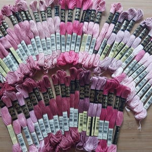 May include: A collection of embroidery floss skeins in various shades of pink, ranging from pale blush to deep magenta. Each skein features a black paper label with white text and a barcode, organized in a fan-like arrangement on a flat surface.