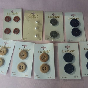 May include: Assortment of La Mode brand buttons in various colors and materials, including wood, plastic, and metal. The buttons are displayed on individual cards with prices and product codes.