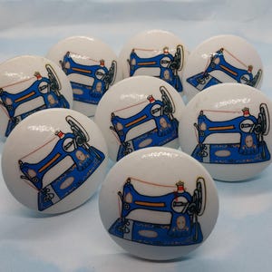May include: Eight white ceramic knobs with blue sewing machine designs. Each knob features a blue sewing machine with red thread and a white background.