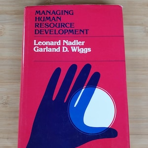 May include: A hardcover book titled "MANAGING HUMAN RESOURCE DEVELOPMENT" by Leonard Nadler and Garland D. Wiggs. The book has a red cover with dark blue graphics of a hand and a white circle.