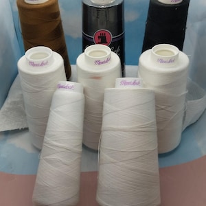 May include: A collection of six spools of white thread with the brand name "Maxi-lock" printed on each spool. The spools are arranged in a semi-circle on a light blue and pink surface.