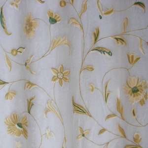 Könnte beinhalten: Nahaufnahme eines weißen Vorhangs mit gestickten Blumenmustern. Das Design zeigt hellgelbe und grüne Blumen, Blätter und Ranken. Die Stickerei verleiht dem Stoff Textur und Details und erzeugt einen eleganten und dekorativen Look.