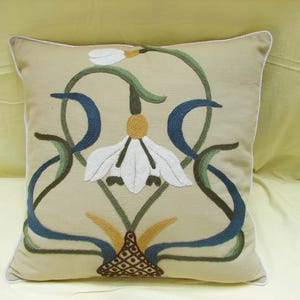 May include: Beige square decorative pillow with an embroidered floral design. The design features white, blue, green, and yellow accents. The pillow has a white trim.