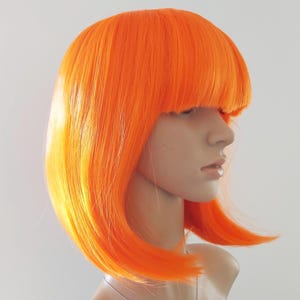 May include: Bright orange wig with a blunt fringe and a bob style. The wig is styled on a mannequin head, showcasing the vibrant color and sleek cut. The hair has a slight gradient of yellow at the ends.