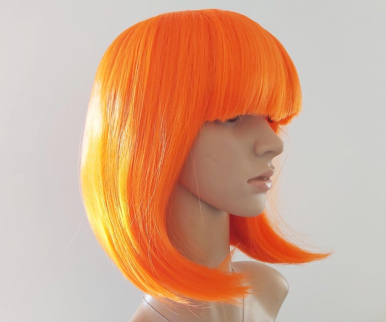 May include: Bright orange wig with a blunt fringe and a bob style. The wig is styled on a mannequin head, showcasing the vibrant color and sleek cut. The hair has a slight gradient of yellow at the ends.