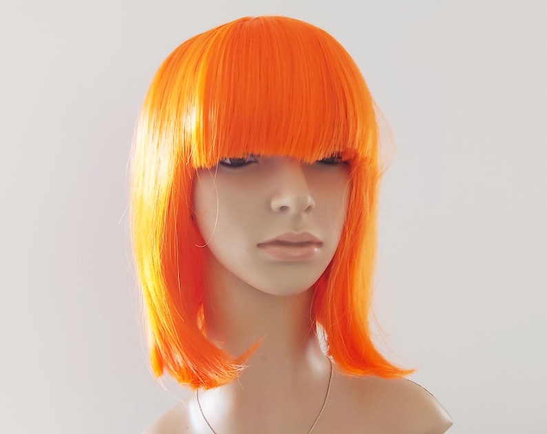 May include: Bright orange wig with a blunt fringe and shoulder-length cut. The wig is styled on a mannequin head, showcasing its vibrant color and sleek design. The wig is a bold fashion statement.