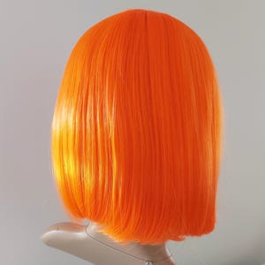 May include: Bright orange wig with a blunt cut, styled in a bob. The wig is displayed on a mannequin head against a plain background. The synthetic hair has a smooth, straight texture and a vibrant, solid color.