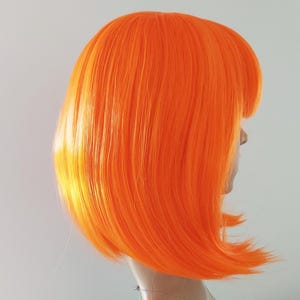 May include: A vibrant orange wig with a blunt bob cut and a side part. The wig is styled on a mannequin head, showcasing the bright, bold color and sleek design. The wig's synthetic fibers appear smooth and shiny.