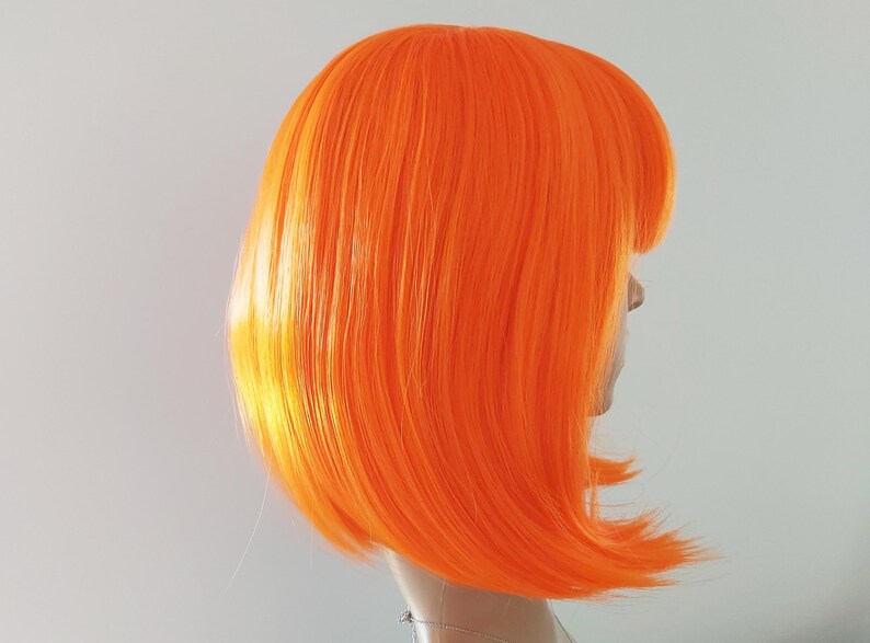 May include: A vibrant orange wig with a blunt bob cut and a side part. The wig is styled on a mannequin head, showcasing the bright, bold color and sleek design. The wig's synthetic fibers appear smooth and shiny.
