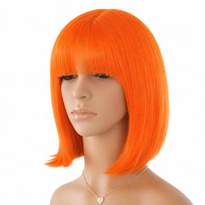 May include: Bright orange bob wig with blunt bangs. The wig is styled with a straight, sleek cut and is designed to sit just below the ears. The wig is on a mannequin head, and a gold necklace with a heart pendant is visible.