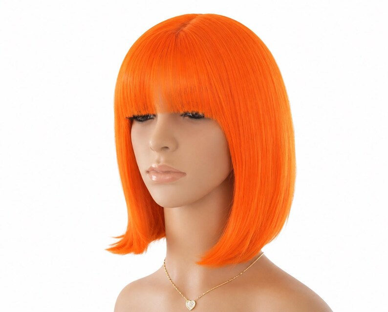 May include: Bright orange bob wig with blunt bangs. The wig is styled with a straight, sleek cut and is designed to sit just below the ears. The wig is on a mannequin head, and a gold necklace with a heart pendant is visible.