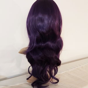 May include: A long, wavy, purple wig with a side part. The wig is styled in loose waves and is made of synthetic hair.