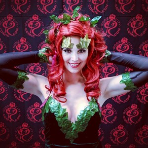 Puede incluir: Una mujer vestida como Poison Ivy, un personaje de DC Comics, con cabello rojo, hojas verdes y un corpi&ntilde;o de terciopelo negro. Est&aacute; posando frente a un fondo negro con un patr&oacute;n repetido de rosas rojas y las palabras "Rose City Comic Con".