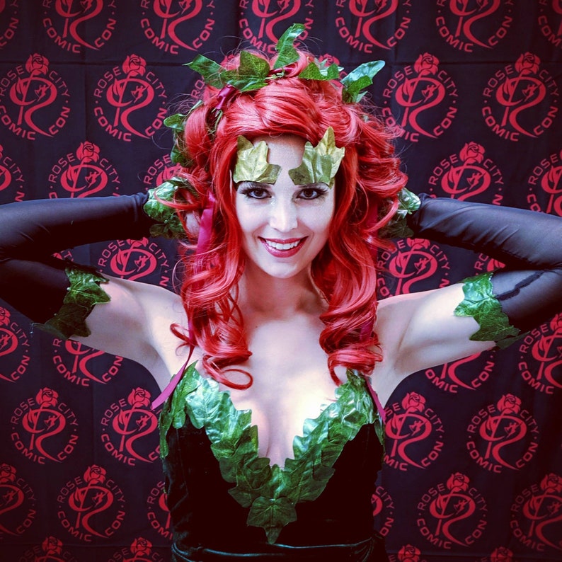 Puede incluir: Una mujer vestida como Poison Ivy, un personaje de DC Comics, con cabello rojo, hojas verdes y un corpi&ntilde;o de terciopelo negro. Est&aacute; posando frente a un fondo negro con un patr&oacute;n repetido de rosas rojas y las palabras "Rose City Comic Con".