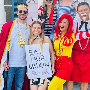 Puede incluir: Un grupo de amigos disfrazados para una fiesta. Una persona lleva un disfraz de rey, otra est&aacute; vestida de vaca, otra de pollo y otra de payaso. Sostienen un cartel que dice "Eat Mor Chikin" con el logotipo de Chick-fil-A.