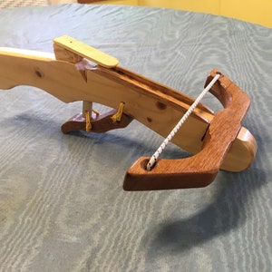 May include: A wooden toy crossbow with a white string and a wooden arrow. The crossbow is made of light and dark wood and has a simple design.