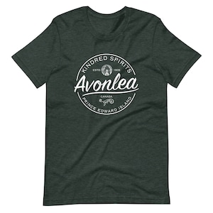 Pode incluir: Uma camiseta verde escura com um logotipo circular branco que diz "Kindred Spirits" com uma árvore e uma casa estilizadas dentro do círculo. O texto "Avonlea" está escrito em itálico abaixo do círculo. O texto "Prince Edward Island" está escrito abaixo do texto em itálico. O texto "Canada" está escrito acima do texto em itálico. O texto "Est. 1909" está escrito dentro do círculo.