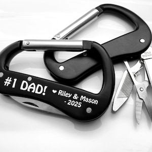 May include: Black carabiner with a multi-tool. The carabiner has the text "#1 DAD!" and the names "Riley & Mason - 2025" in white. The multi-tool includes a knife, scissors, and a file. The carabiner is a practical gift.