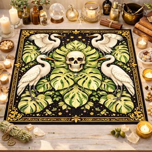 May include: A black decorative mat featuring a central skull surrounded by large green leaves and four white herons. The mat has a gold border and is decorated with small stars and floral elements. The overall design has a mystical or gothic theme.