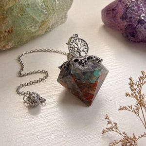 May include: A multi-coloured crystal pendulum with a silver chain and a decorative tree of life charm. The crystal has red, green, and blue hues. Two large crystals are in the background.