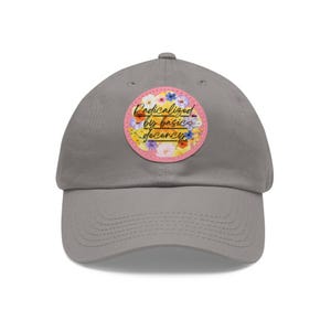 May include: A gray baseball cap with a curved brim. The cap features a circular patch with the text "Radicalized by basic decency" in a script font, surrounded by a floral design with pink, yellow, and blue flowers.