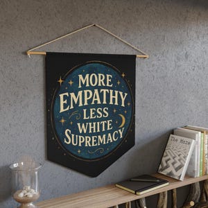 May include: A black banner with the text "MORE EMPATHY LESS WHITE SUPREMACY" in a circular design with stars and a crescent moon. The banner is suspended from a wooden rod with a string.