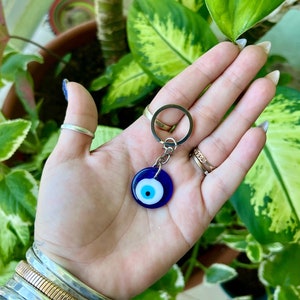 May include: A silver key chain with a blue and white evil eye charm. The charm is round and has a white pupil and a blue iris.
