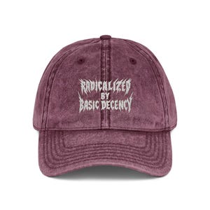 May include: A maroon baseball cap with a curved peak. The cap displays the words "RADICALIZED BY BASIC DECENCY" in white, stylized lettering. The hat has a vintage, worn appearance.