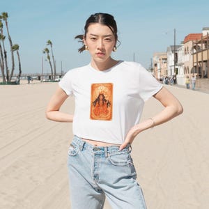 May include: White crop top with a tarot card graphic in shades of orange and gold. The graphic features a woman with long hair and a crown. The model is wearing light wash blue jeans and is standing on a sandy beach.