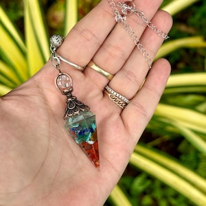 May include: A clear, cone-shaped pendulum with colourful stones encased in resin. The pendulum has a silver chain and a decorative metal cap. The stones include red, blue, green, and white. The pendulum is designed for spiritual practices.