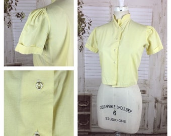 May include: A pale yellow short-sleeved button-up shirt with a mandarin collar. The shirt has a slightly cropped fit and is made of a lightweight fabric.