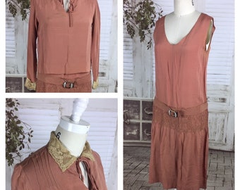 Pode incluir: Um vestido vintage dos anos 1920 de estilo flapper, na cor rosa poeira, com colar de renda e cinto. O vestido tem um corpete sem mangas e uma saia com cintura franzida.