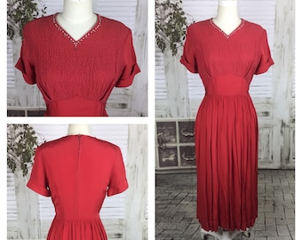 Pode incluir: Um vestido vintage vermelho com um corpete justo e uma saia cheia. O vestido tem um decote em V e mangas curtas. O decote é adornado com uma fileira de strass.