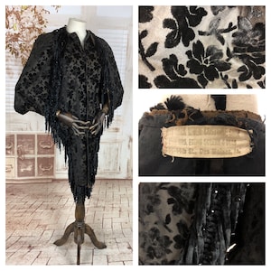 Op de afbeelding: Een zwarte fluwelen cape met een bloemenpatroon en kralenfranjes. De cape wordt getoond op een mannequin en heeft een label met de tekst "Black Velvet Evening Cape,  U.S.  Empire  Co.,  San  Francisco,  Cal."