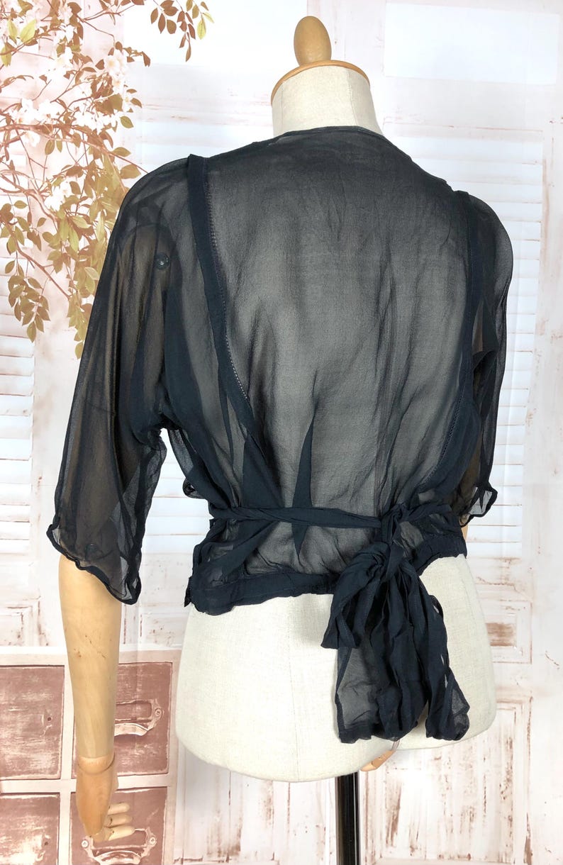 May include: Black sheer wrap top with short sleeves and a tie closure at the waist. The top is displayed on a mannequin, showcasing its delicate, flowing fabric. The garment's design is simple yet elegant, suitable for layering or as a standalone piece.