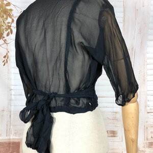 May include: Black sheer blouse with three-quarter sleeves and a tie-back detail. The blouse is displayed on a mannequin, showcasing its delicate fabric and design. The top has a button closure on the sleeve.