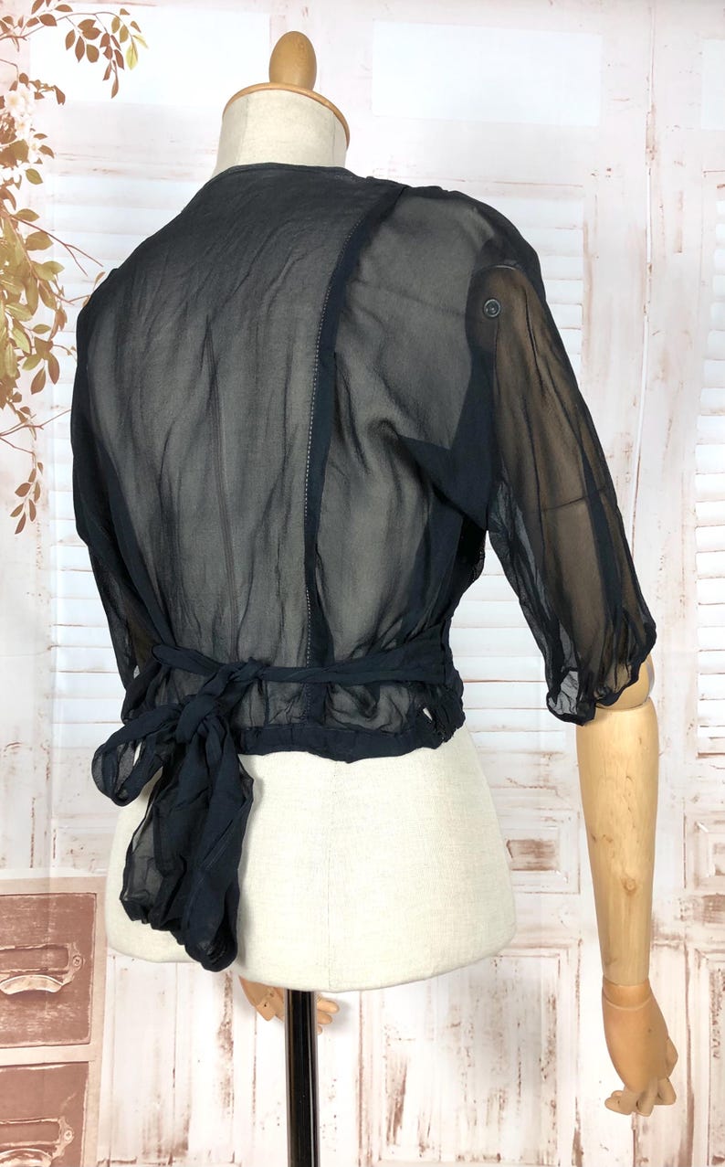 May include: Black sheer blouse with three-quarter sleeves and a tie-back detail. The blouse is displayed on a mannequin, showcasing its delicate fabric and design. The top has a button closure on the sleeve.