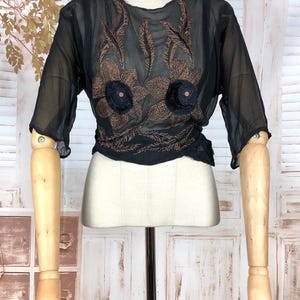 May include: Black sheer blouse with short sleeves and intricate brown floral embroidery. The blouse is displayed on a mannequin with wooden arms and a cream-colored torso, set against a white background.