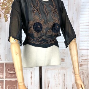 May include: Black sheer blouse with brown floral embroidery and two black flower accents. The blouse has three-quarter sleeves and a cinched waist. The garment is displayed on a mannequin.