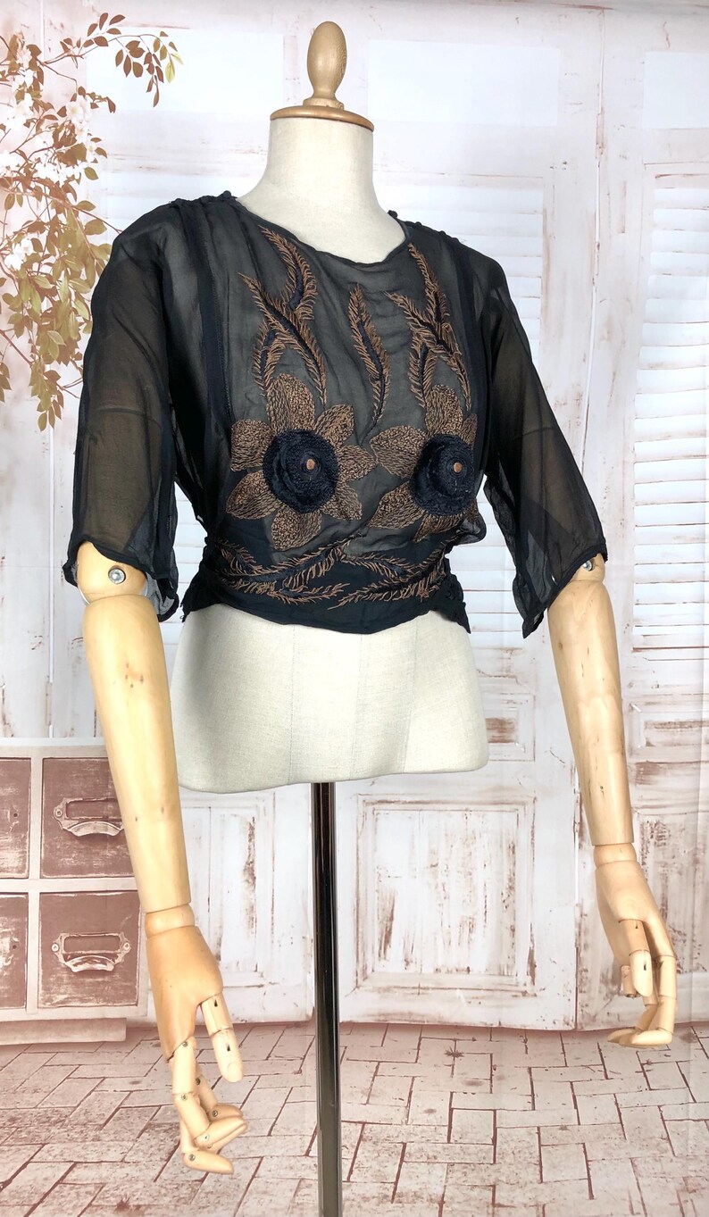 May include: Black sheer blouse with brown floral embroidery and two black flower accents. The blouse has three-quarter sleeves and a cinched waist. The garment is displayed on a mannequin.