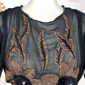 May include: A sheer, dark navy blue blouse with intricate embroidery. The design features bronze-colored feather and floral patterns, with navy blue accents. The blouse has a round neckline and long sleeves, offering a vintage aesthetic.