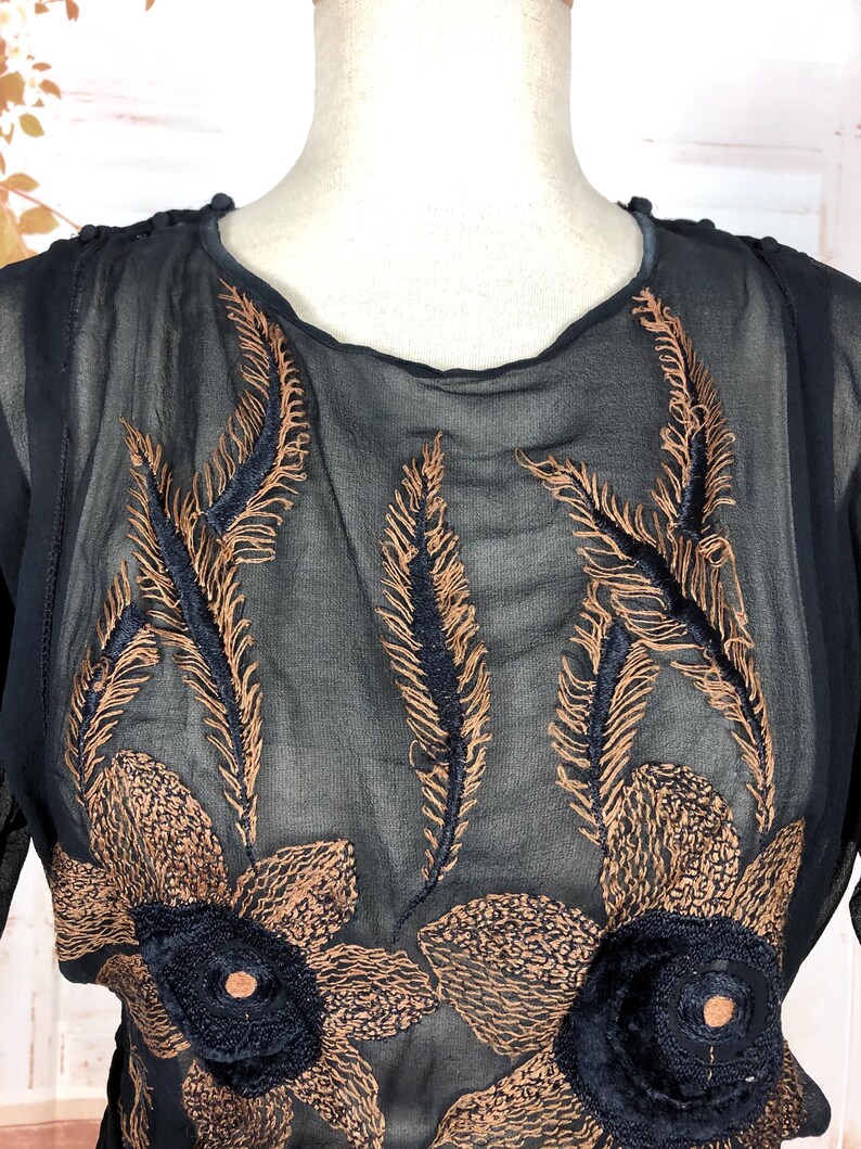 May include: A sheer, dark navy blue blouse with intricate embroidery. The design features bronze-colored feather and floral patterns, with navy blue accents. The blouse has a round neckline and long sleeves, offering a vintage aesthetic.