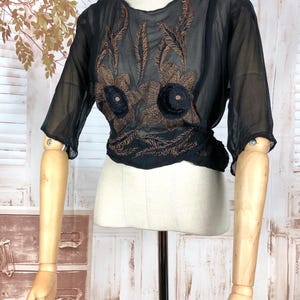 May include: Black sheer blouse with brown floral embroidery. The blouse features short sleeves and a gathered waist. The embroidery is concentrated on the bodice, creating a decorative focal point. The blouse is displayed on a mannequin.