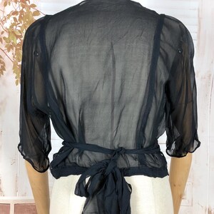 May include: Black sheer bolero top with short sleeves and a tie closure at the waist. The bolero is displayed on a mannequin, showcasing its delicate, flowing fabric and design. The top has a ruffled hem and is made of a lightweight material.